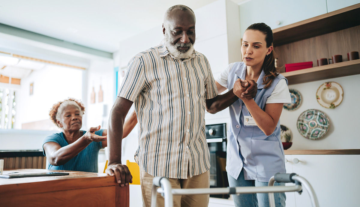 Wife sitting helping husband up from seat while female nurse helps him to walker